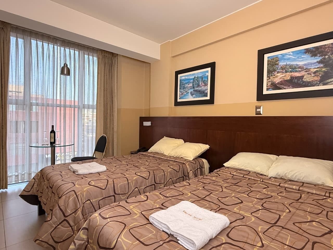 Quarto de hotel com duas camas, uma mesinha com uma garrafa e copos, e janelas amplas com cortinas.
