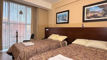 A hotel room with two beds, a small table with a bottle and glasses, and large windows with curtains.