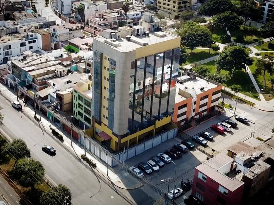Vista aérea de un edificio de varios pisos con fachada central de vidrio, rodeado de edificios más pequeños y un estacionamiento.