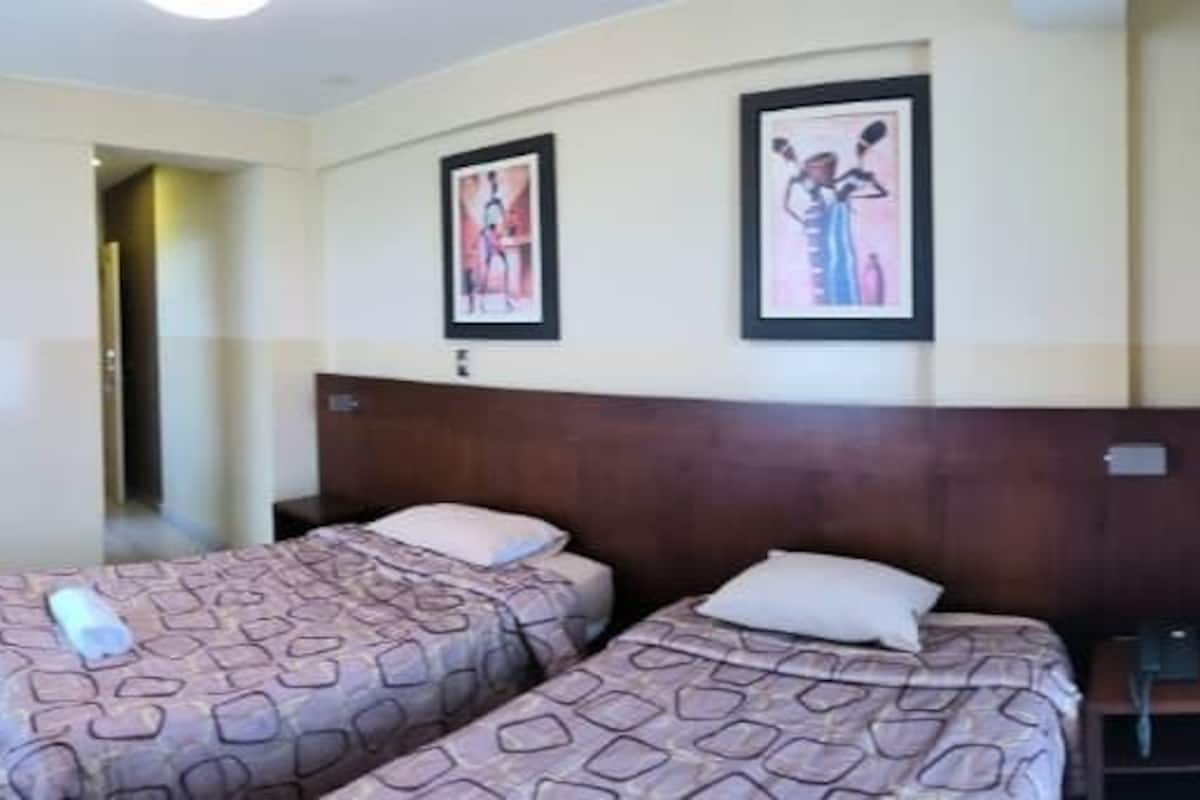 A hotel room with three beds, a TV, a heater, and two framed pictures on the wall.