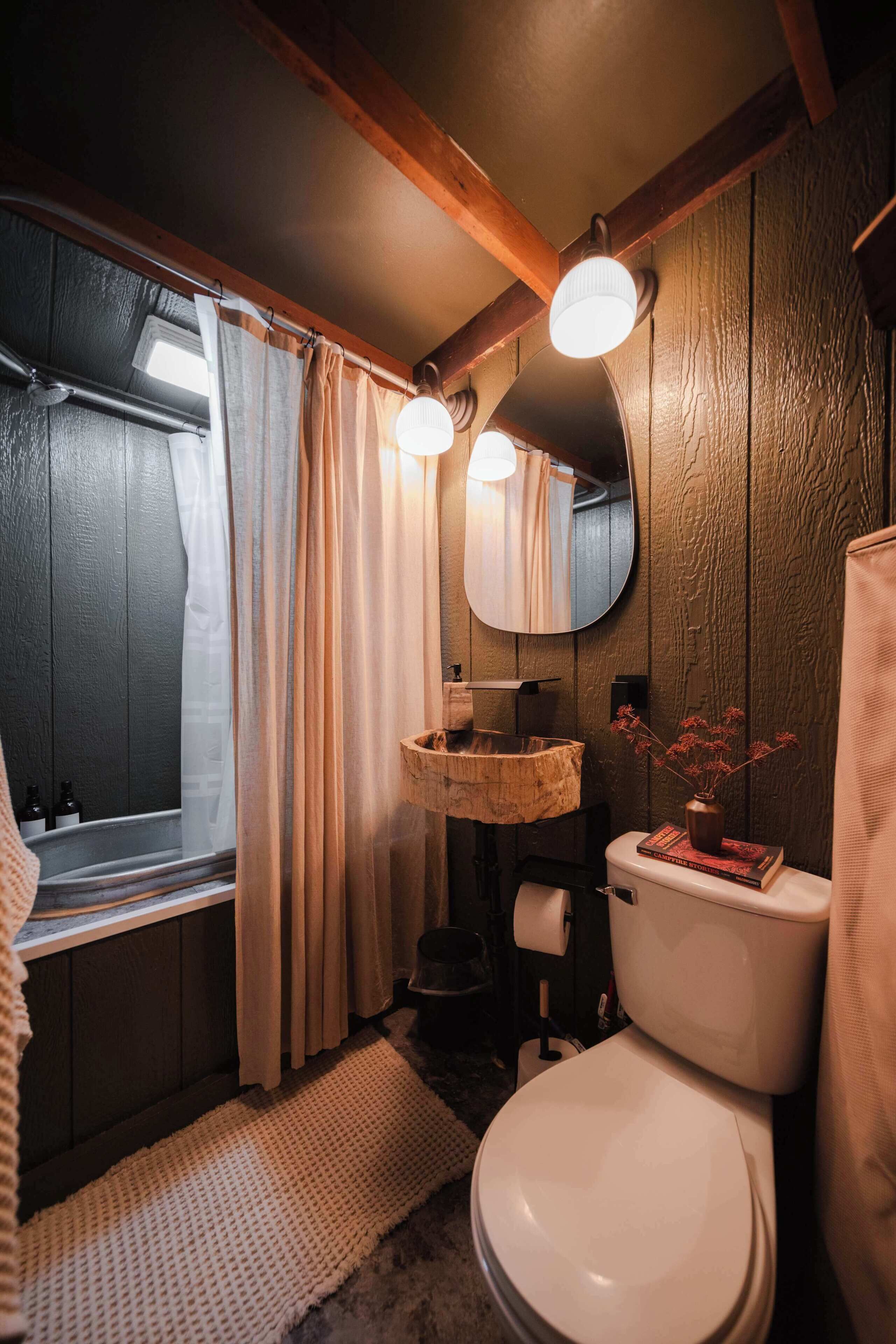 Un baño con paredes de madera, una ventana con cortinas, un lavamanos con cuenca de madera, un inodoro y un espejo.