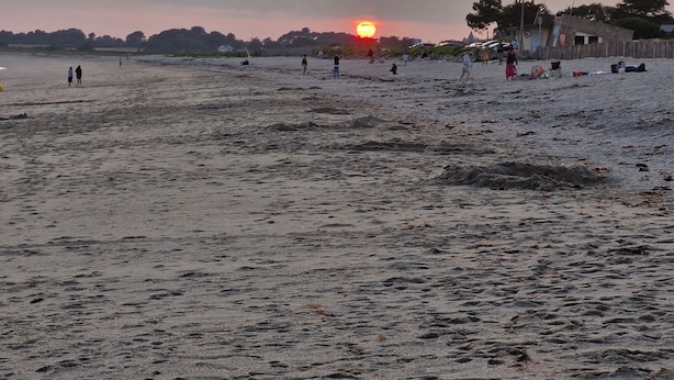 Een strand met mensen die wandelen en zitten, een ondergaande zon op de achtergrond en enkele gebouwen in de verte.