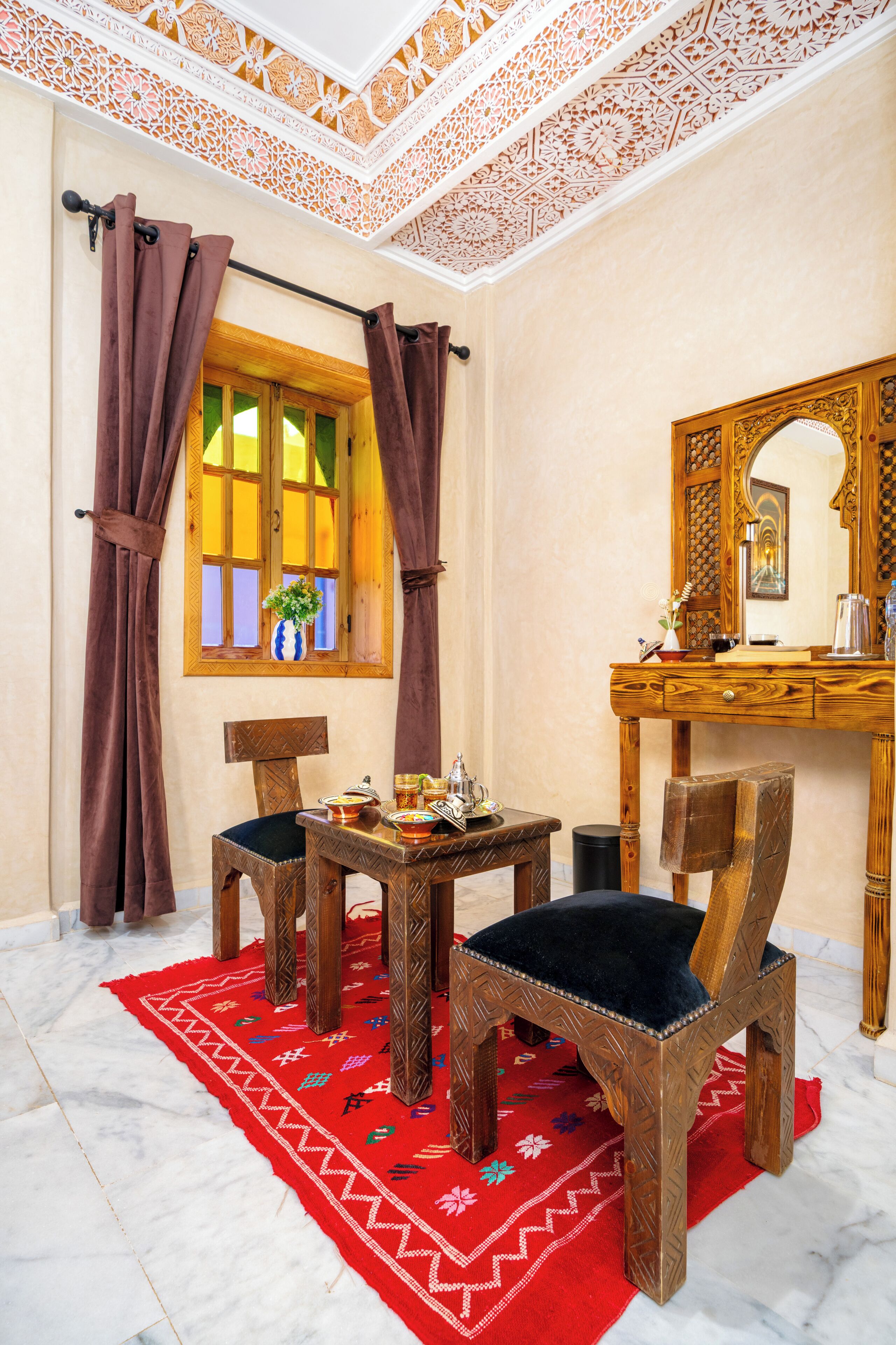 A room with a wooden desk, two chairs, a red rug, a window with curtains, and a decorative ceiling.