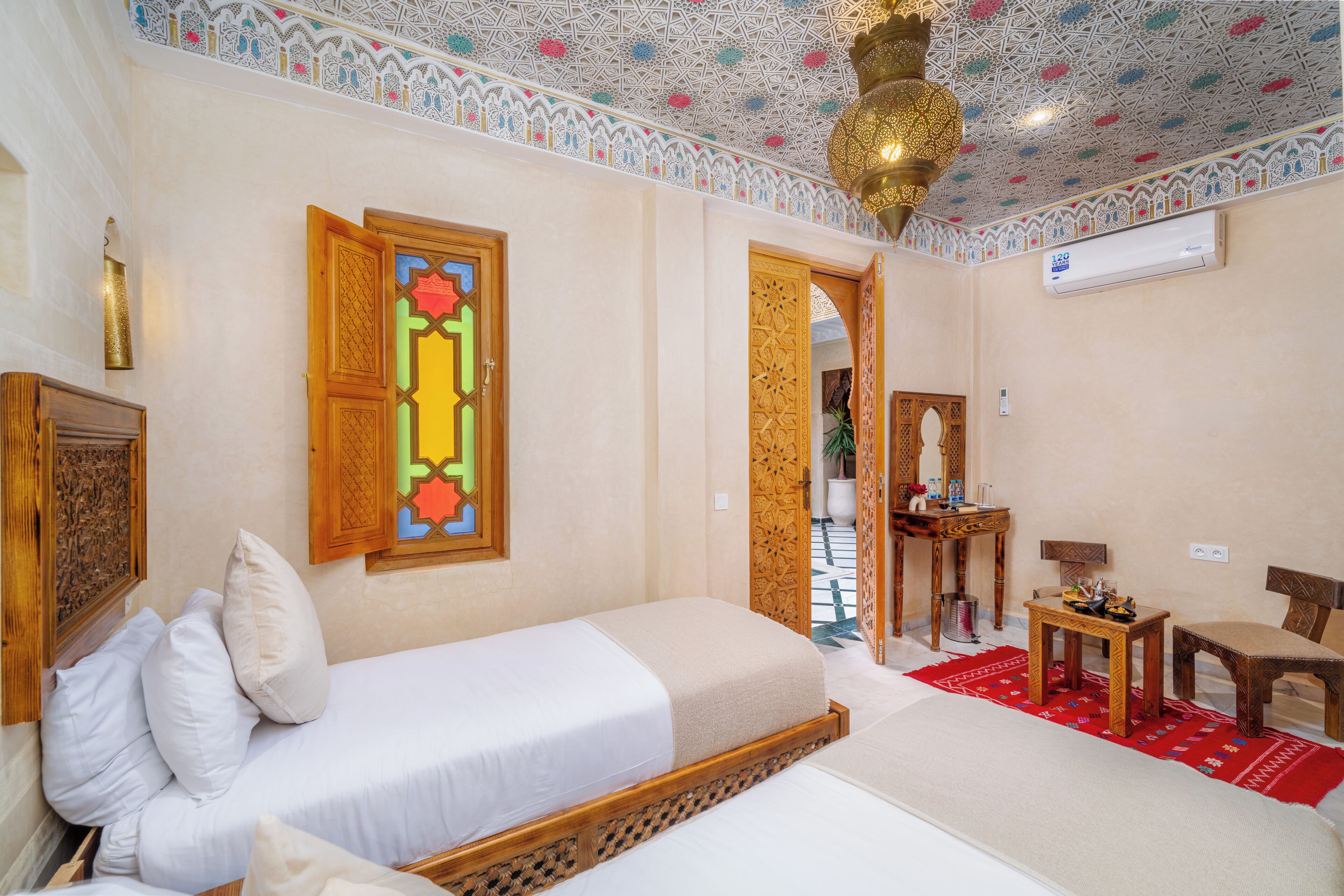 A room with two beds, a wooden door, a red patterned rug, a small table, and a ceiling with intricate tile work.