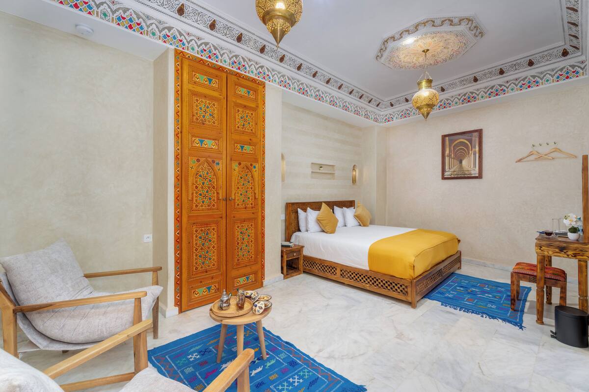 A room with a bed, a chair, a table with a teapot, a hanging plant, and a decorative ceiling.