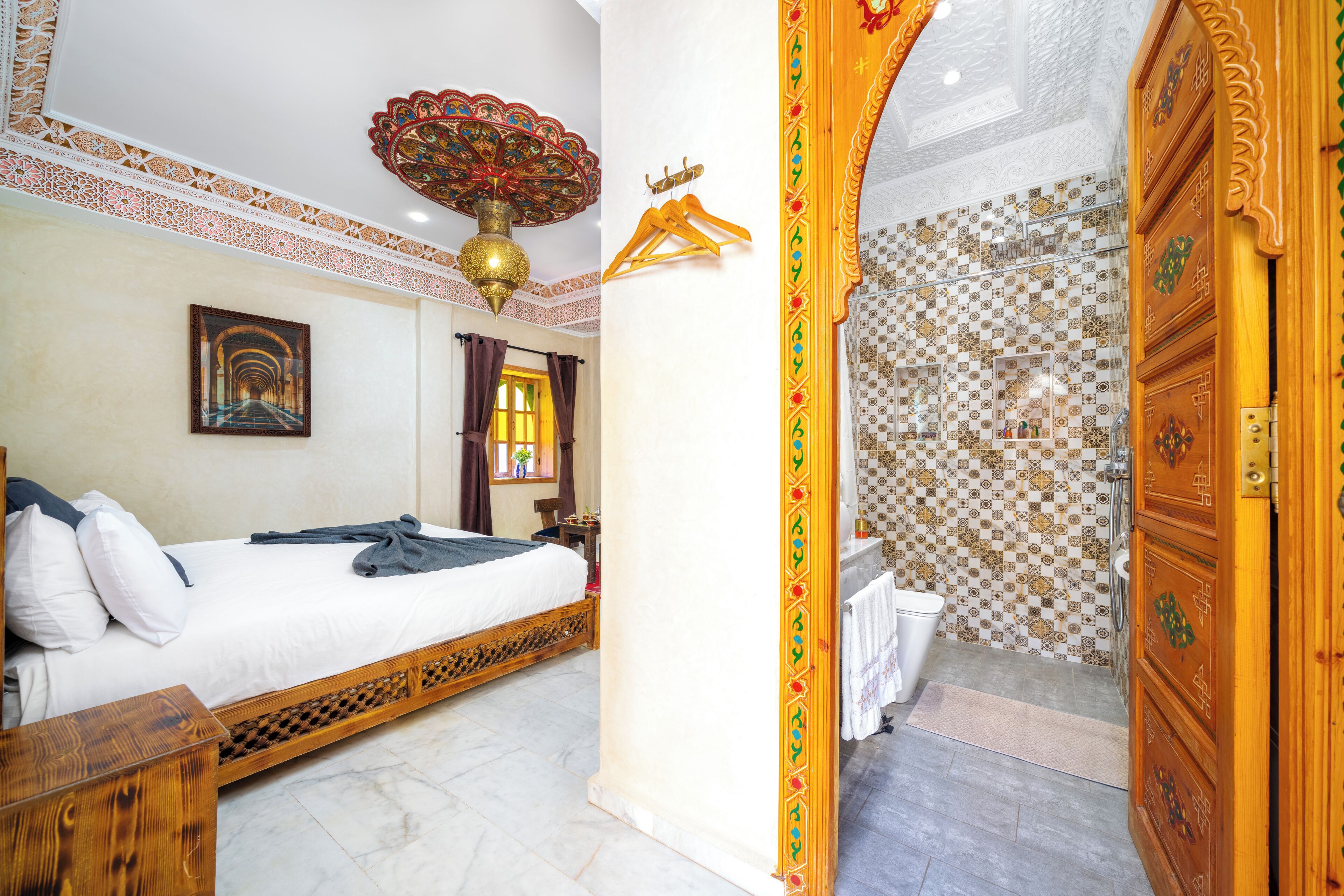 A bedroom with a bed, a wall-mounted lamp, a picture, and a bathroom with a tiled wall and a wooden door.