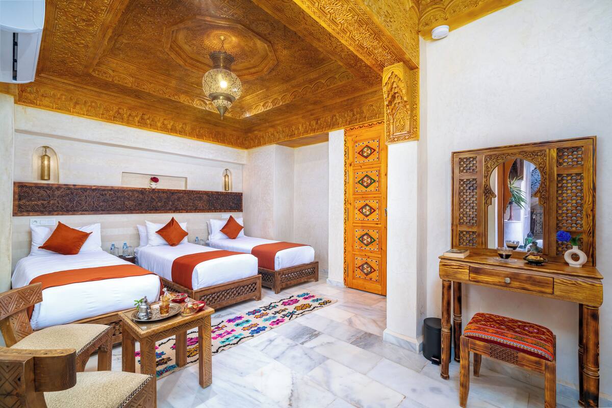 A hotel room with two beds, a wooden desk, a chair, a small table, and a decorative mirror.