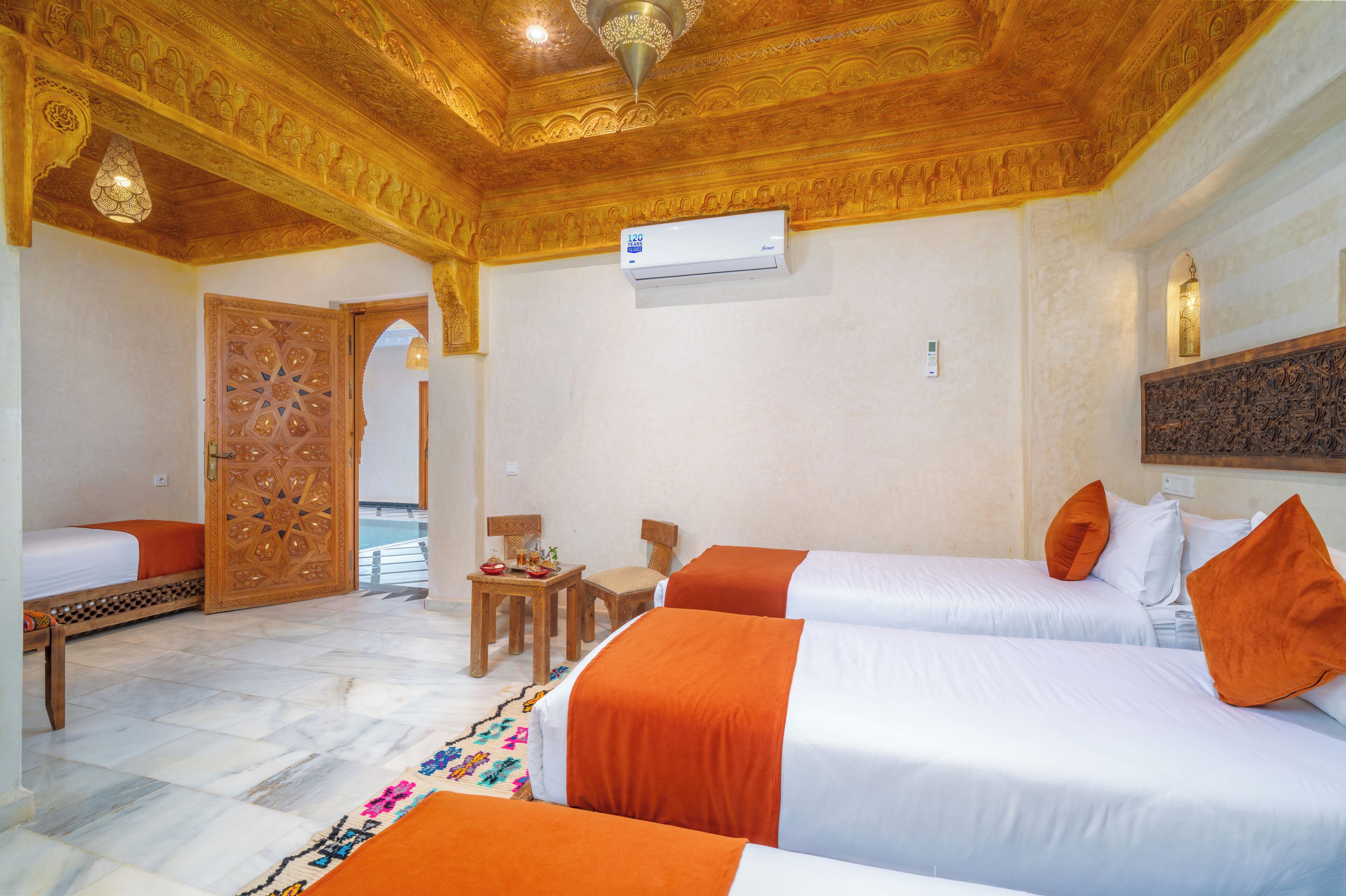 A hotel room with two beds, a wooden door, a small table with a chair, and an air conditioning unit.