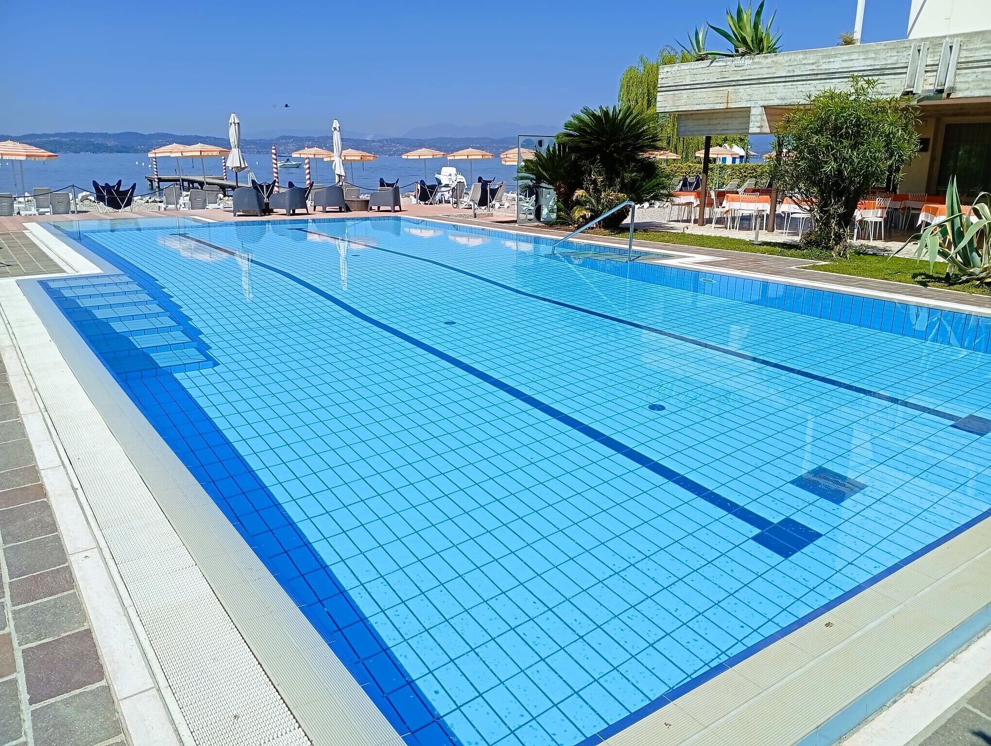 Ein klares, blaues Schwimmbecken mit einer gefliesten Liegefläche, Liegestühlen, Sonnenschirmen und Meerblick.