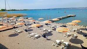 A beach with sun loungers and striped umbrellas, a pier extending into the water, and boats in the background. - Hotel Tre Vele (Sirmione)