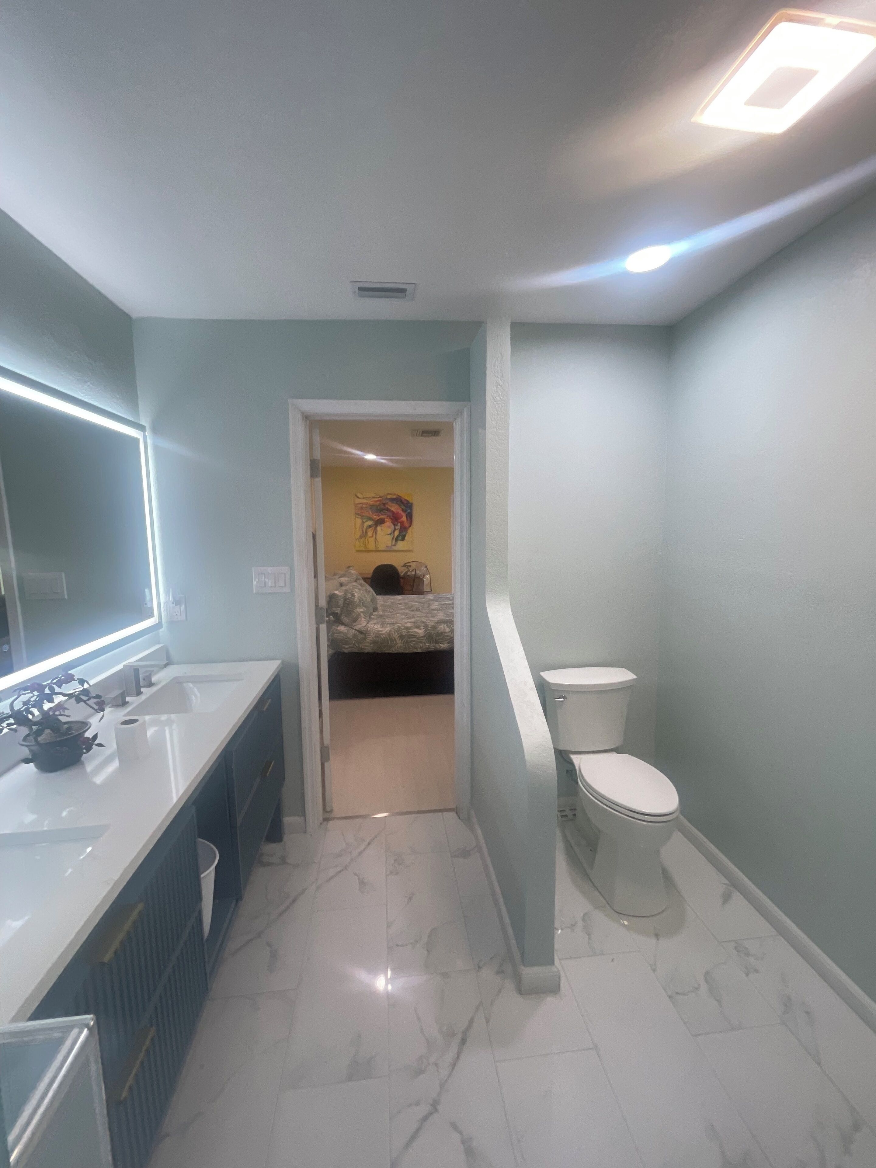 Ein Badezimmer mit Waschbecken, Spiegel und Toilette. Eine Tür führt zu einem Schlafzimmer mit Bett und einem farbenfrohen Bild an der Wand.
