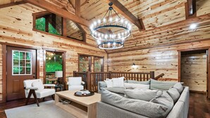 A log cabin interior with a wooden ceiling, a chandelier, a gray sofa, a white chair, and a wooden dining table. - Big Chief Lodge (Sevierville)