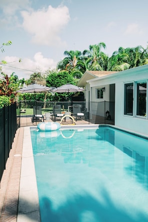 Ein Swimmingpool mit Liegestühlen und Sonnenschirmen, umgeben von einem Zaun und Palmen.