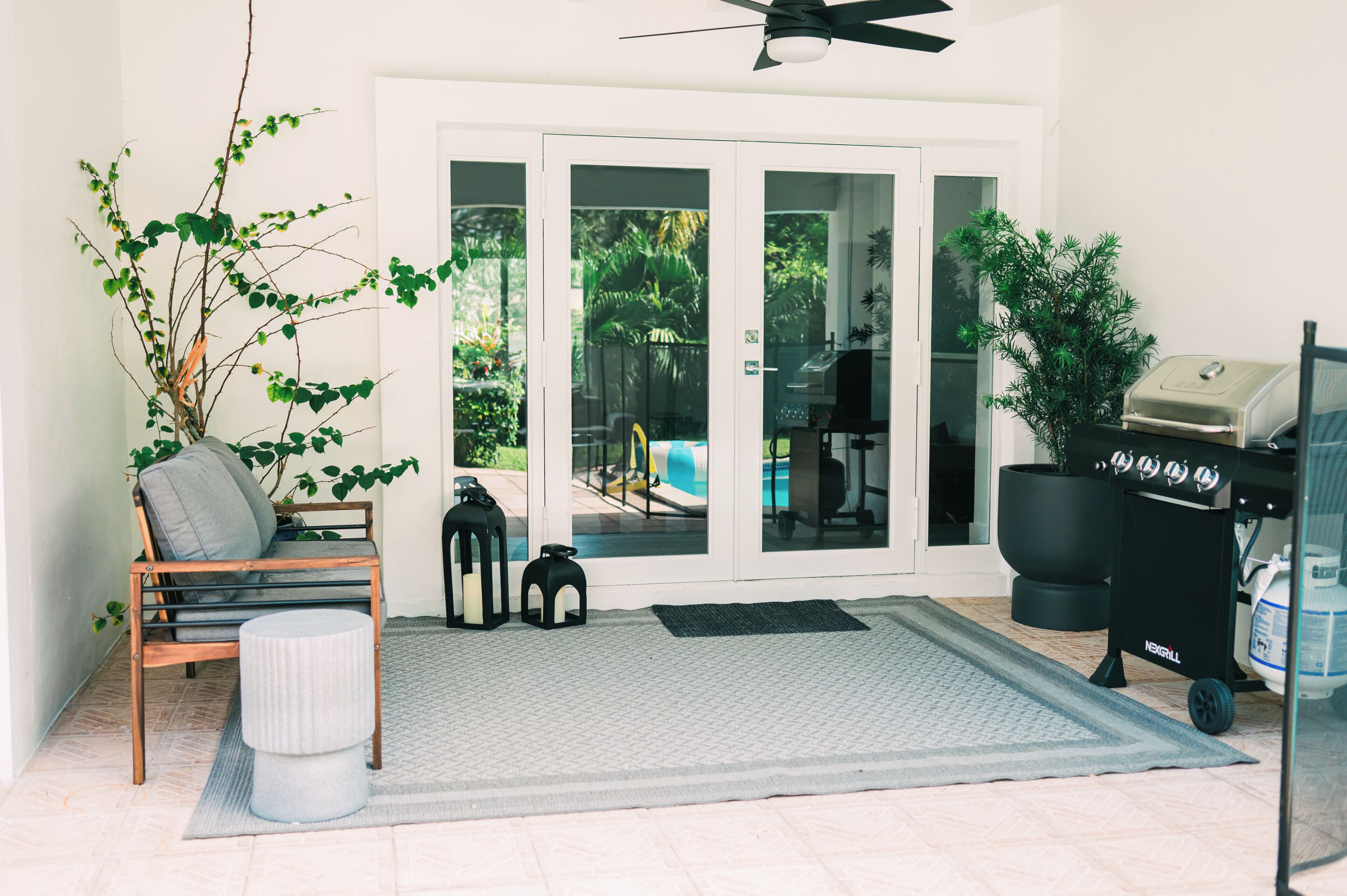 A patio area with a grill, a chair, a small table, a potted plant, and a view of a swimming pool through glass doors.