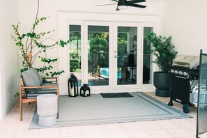 Eine Terrasse mit Grill, einem Stuhl, einem kleinen Tisch, einem Blumentopf und Blick auf ein Schwimmbecken durch Glasüren.