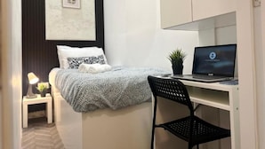 Une chambre à coucher comprenant un lit, un bureau avec un ordinateur portable, une chaise, une petite table avec une lampe et une étagère fixée au mur.