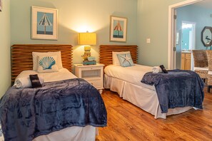 A room with two beds, each with a wooden headboard and a blue blanket. There is a nightstand with a lamp and a decorative item between the beds. The walls are painted light blue, and there are framed pictures above the beds. - The Rookery III 4009 (Gulf Shores)