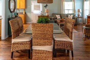 A dining area with wicker chairs and a glass table, surrounded by wooden flooring and a view of the living room.