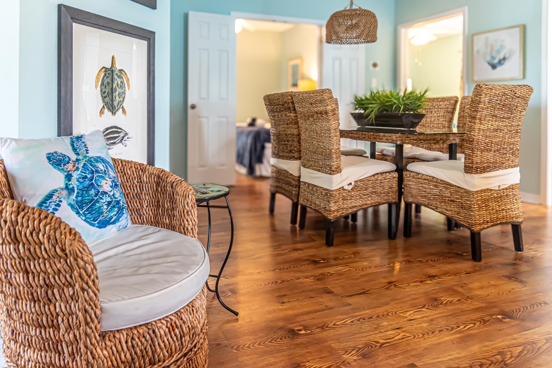 A dining area with wicker chairs and a glass table, framed artwork on the wall, and a wooden floor.