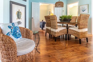 A dining area with wicker chairs and a glass table, framed artwork on the wall, and a wooden floor.