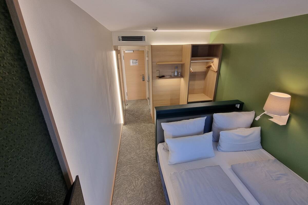 A hotel room with a bed, a bedside table, a lamp, a wardrobe, and a bathroom visible through an open door.