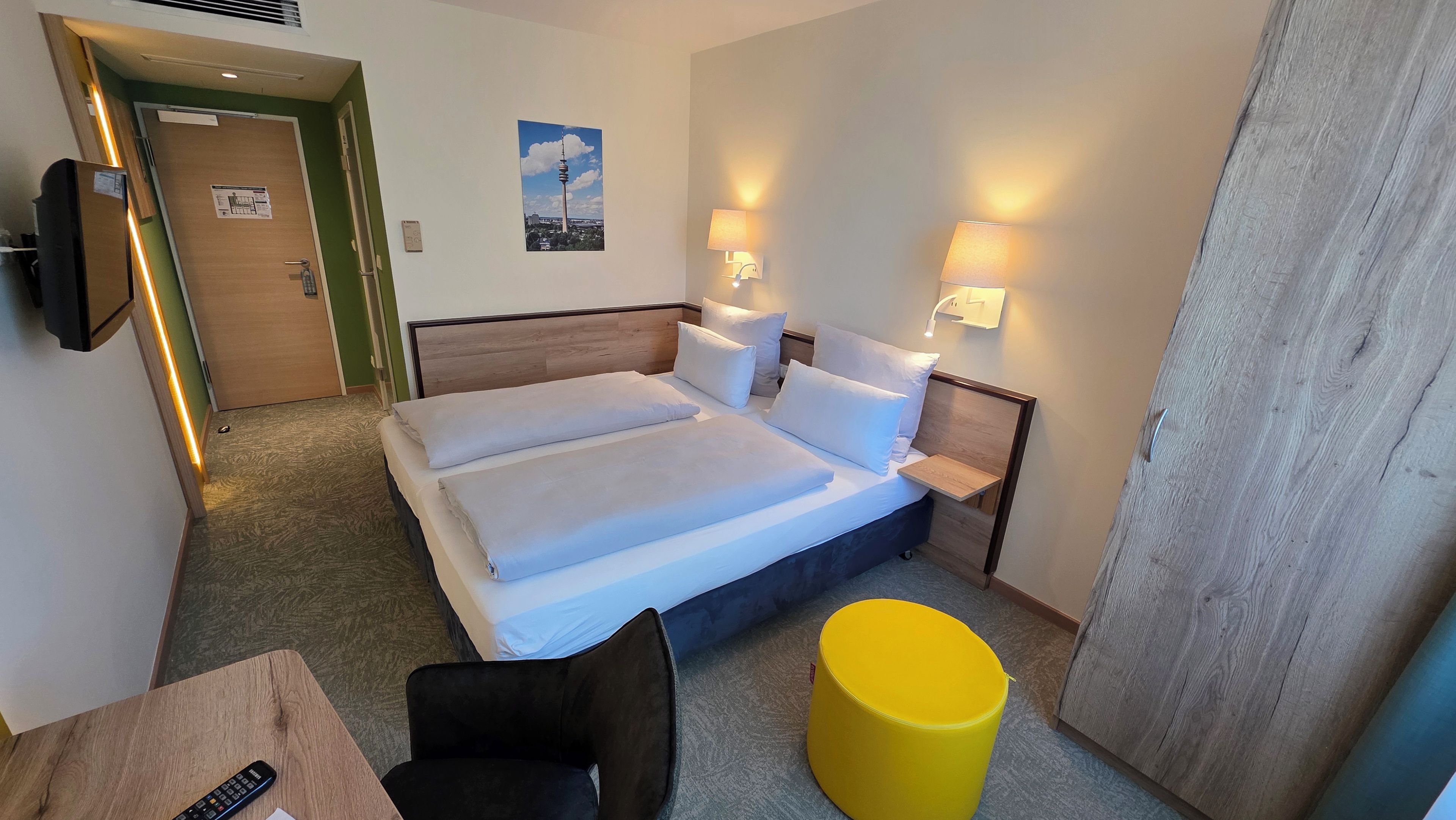 A hotel room with a double bed, bedside tables, a chair, a small table, and a yellow ottoman.