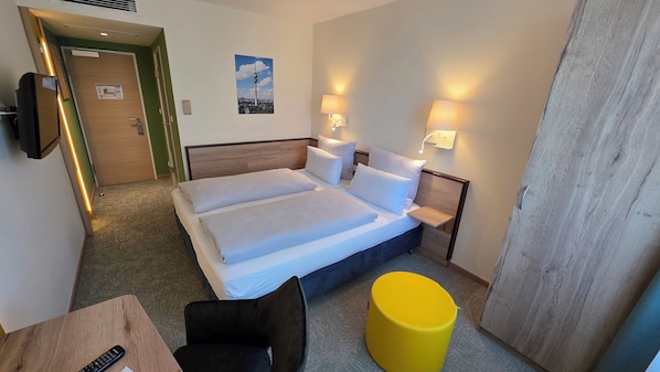 A hotel room with a double bed, bedside tables, a chair, a small table, and a yellow ottoman. - my MINGA City Sendlinger Tor (Munich)