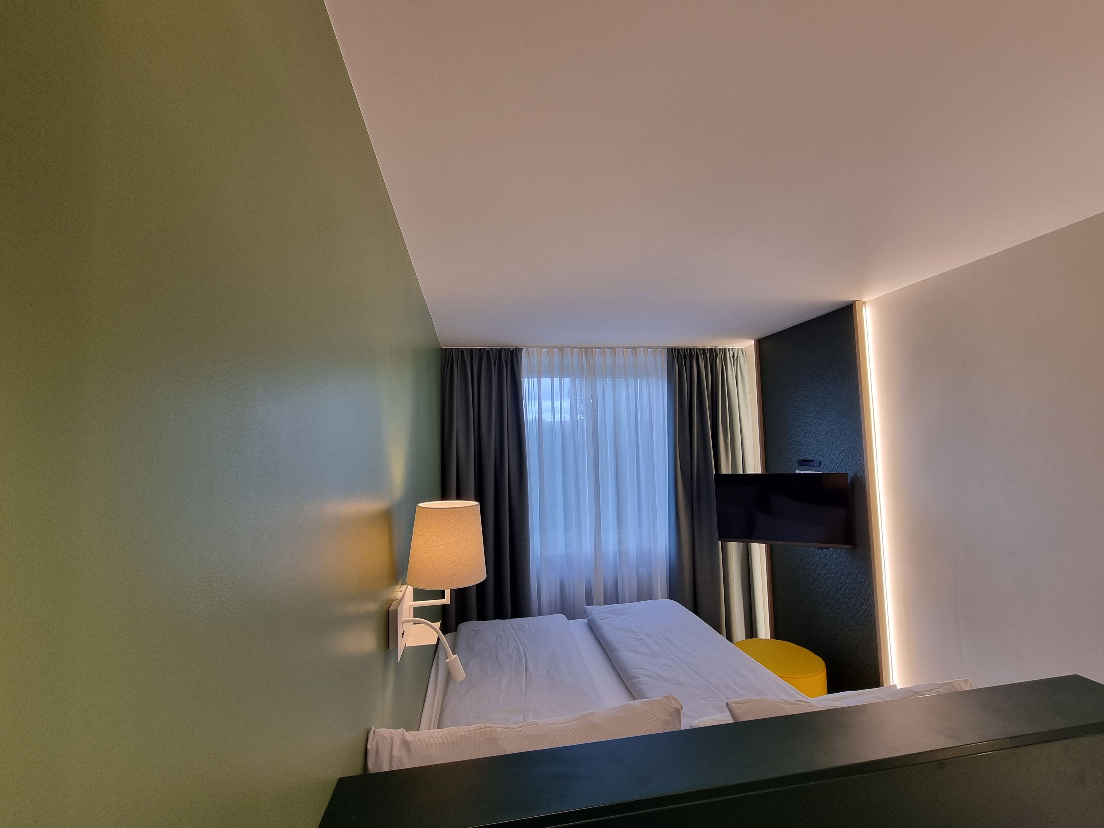 Une chambre d’hôtel avec un lit, une lampe de chevet, un téléviseur fixé au mur et une fenêtre avec des rideaux.
