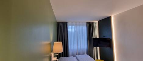 Une chambre d’hôtel avec un lit, une lampe de chevet, un téléviseur fixé au mur et une fenêtre avec des rideaux.