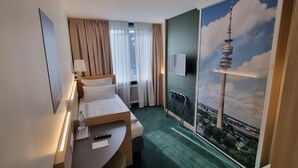 Hotelkamer met een bed, bureau, lamp, raam met gordijnen en uitzicht op een stadsbeeld.