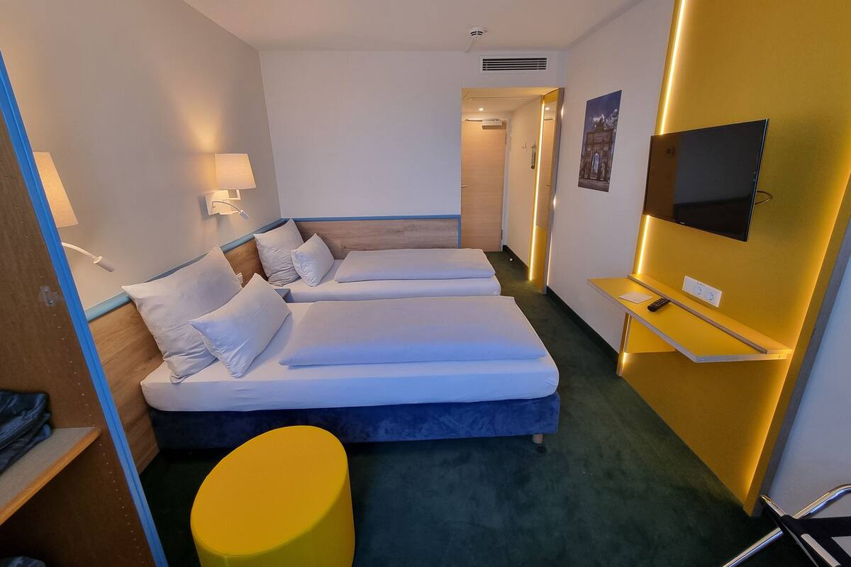 A hotel room with two beds, a TV, a desk, and a yellow ottoman.