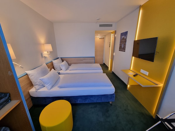 A hotel room with two beds, a TV, a desk, and a yellow ottoman. - my MINGA City Sendlinger Tor (Munich)