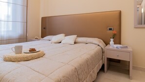 A neatly made bed with a beige headboard, a bedside table with a book and a vase, and a window with sheer curtains. - Elegant Sea View Apartment with Panoramic Patio (Grottammare)