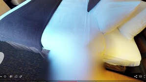 Hotellihuone, jossa on sänky, yöpöytä, lamppu ja ovi, joka johtaa käytävään.