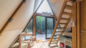 Ein quadratisches Zimmer im Dachgeschoss mit Holztisch, Stühlen, einer Leiter und einer Schiebetür, die auf eine Terrasse im Freien führt.