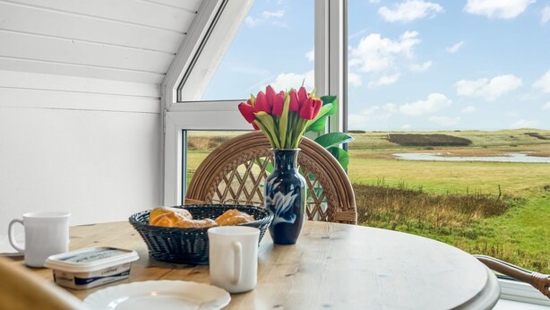 Une table avec des aliments pour le petit-déjeuner, un vase rempli de fleurs et, par la fenêtre, une vue sur un paysage verdoyant.