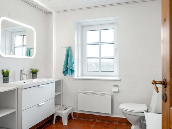 A modern bathroom with a white sink, mirror, and toilet. There is a window with a view, a radiator, and a small stool.