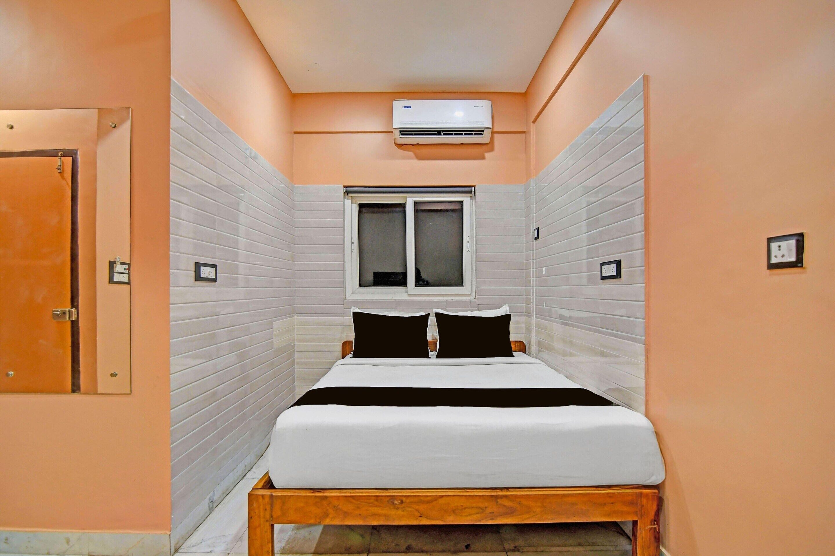 A narrow hotel room with a bed, a window, an air conditioner, and a door.