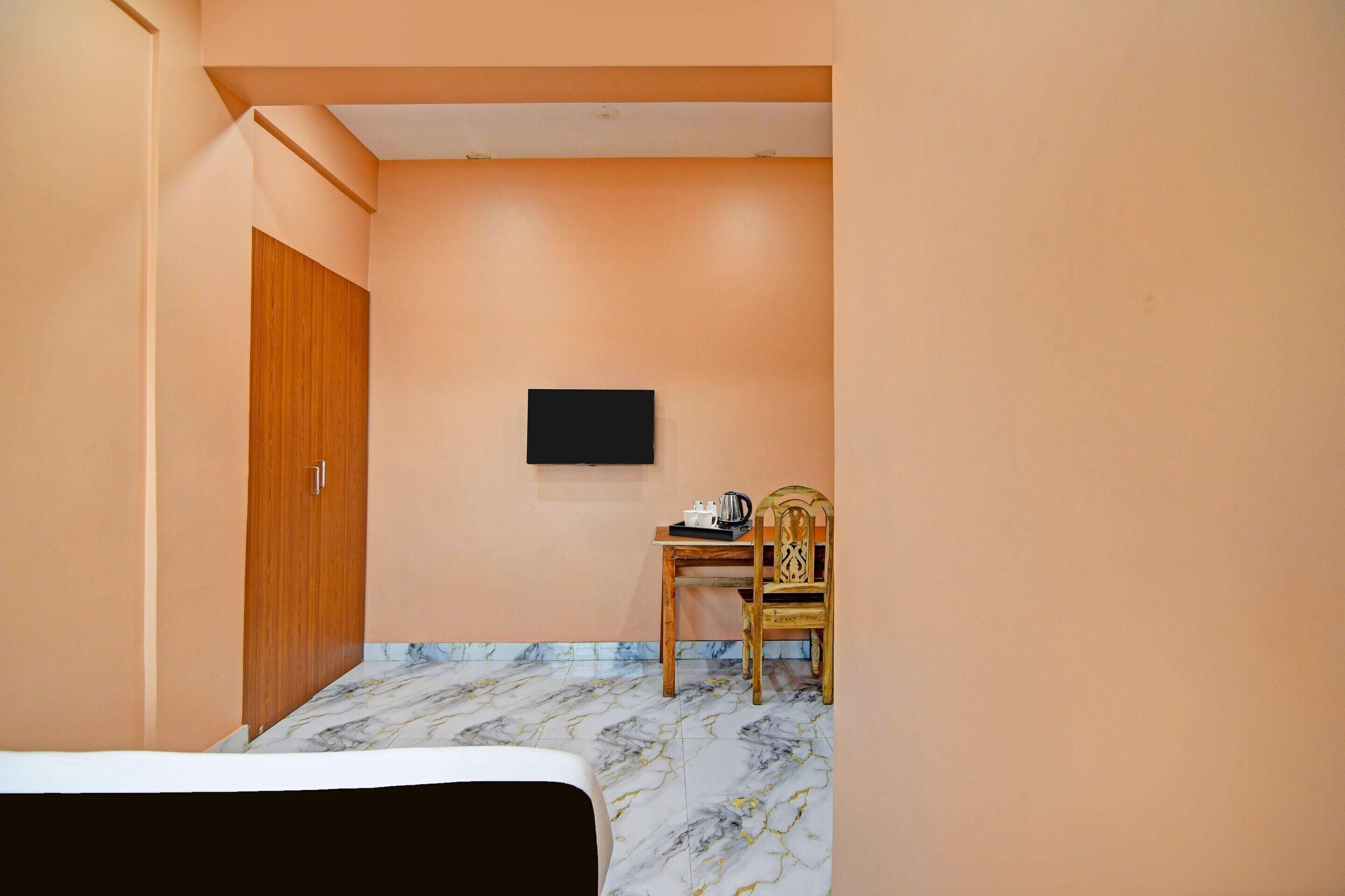 Ein Zimmer mit einem Bett, einer Holztür, einem an der Wand befestigten Fernseher, einem Holzstuhl und einem Tisch mit einem Wasserkocher.