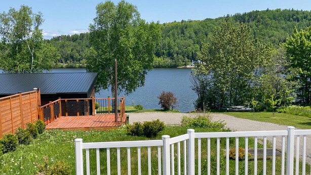 A lakeside property with a wooden deck, white fence, and a view of a lake and forested hills. - Nature, Pool Table & Water Access | Le Highland (Saint-Félix-d'Otis)