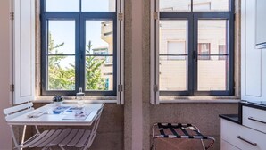 Ein Zimmer mit Tisch, einer Bank, einem Fenster mit Blick auf Gebäude und einem kleinen Hocker.