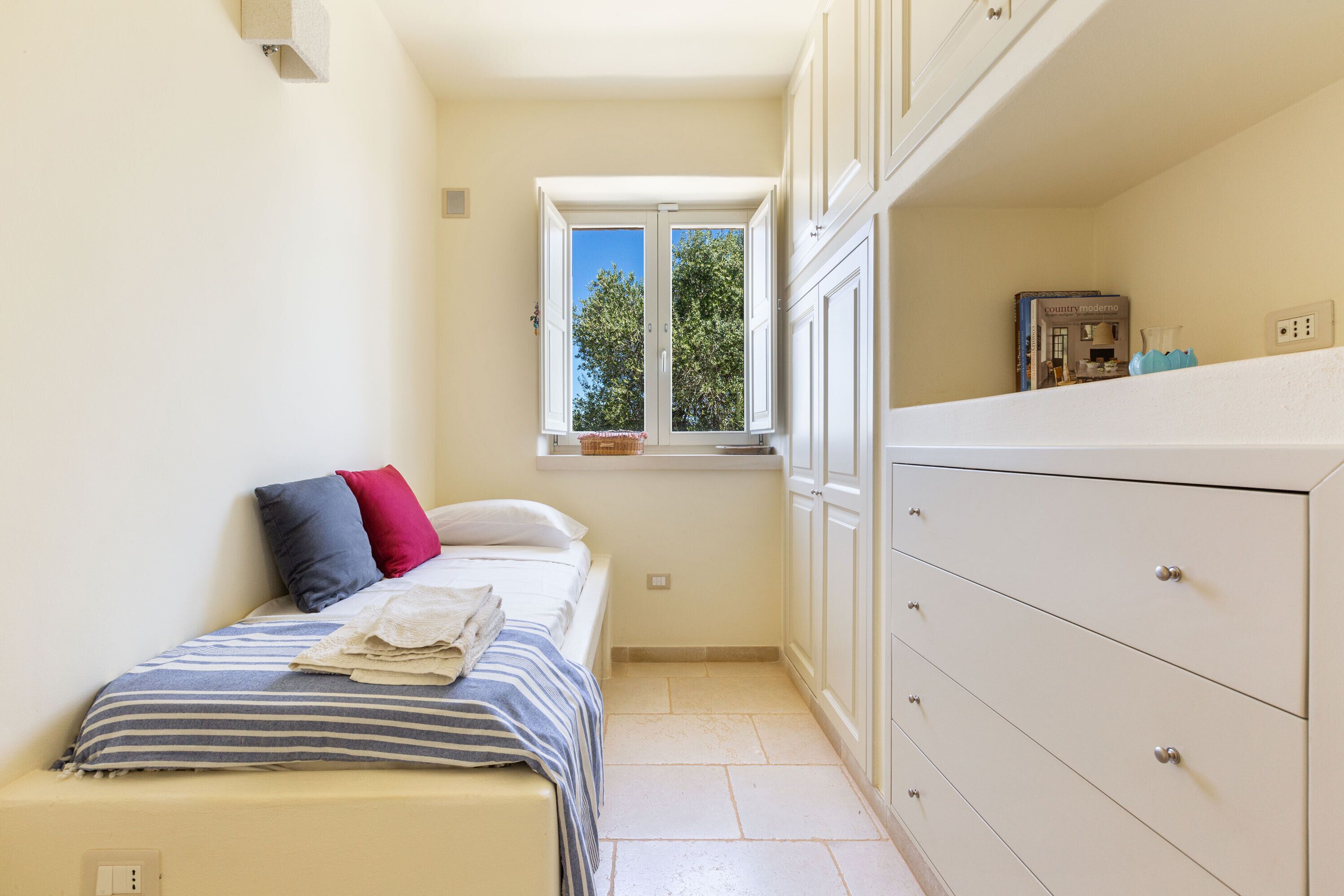 Ein kleines, gemütliches Schlafzimmer mit einem Einzelbett, einem Fenster mit Blick auf Bäume und einer weißen Kommode mit Schubladen.