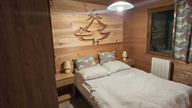 Ett sovrum i en stuga med träpanel, inrett med en säng, två sängbord, en lampa, ett element och ett fönster med gardiner.