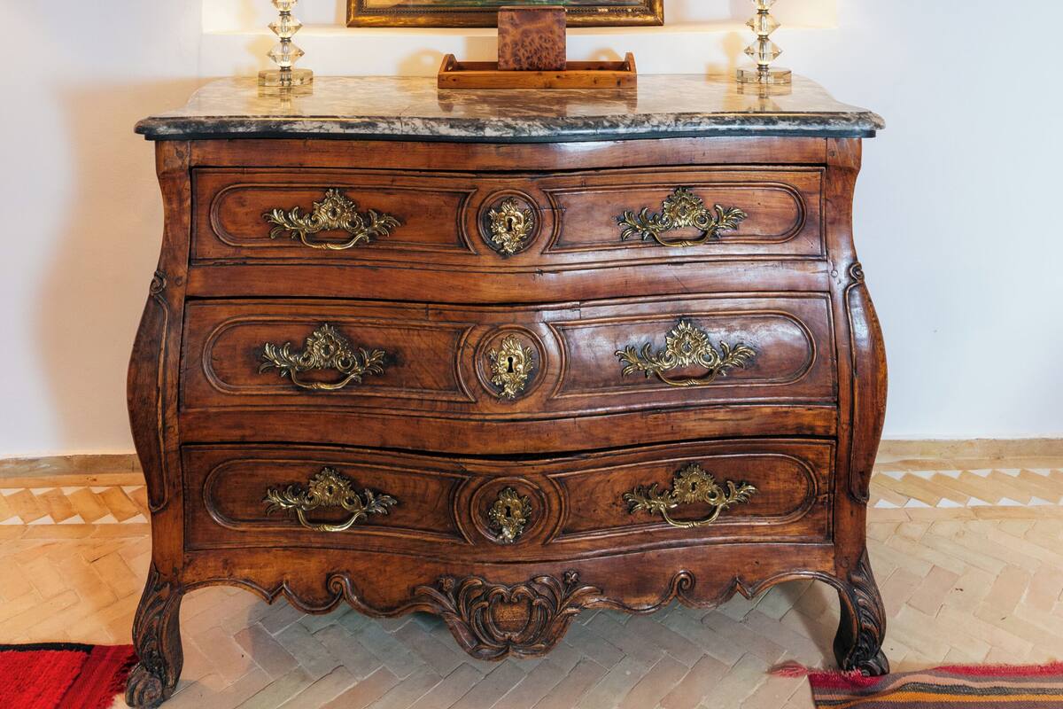 A wooden dresser with ornate handles, a painting above it, and two lamps on either side.