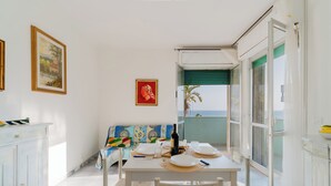 Uma área de refeições com mesa posta para quatro pessoas, um sofá e vista para o mar através de uma porta deslizante.