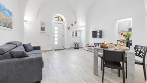 A modern living room with a grey sofa, a dining table with chairs, a flat-screen TV, and a kitchen area with a window. - Dimora Aurea by BarbarHouse (Oria)