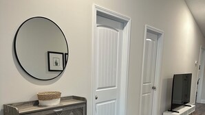 A hallway with a herringbone-patterned dresser, a round mirror, and a framed picture on the wall. There are white doors and a television on a stand. - 2-bedroom townhome in South Augusta (Augusta)