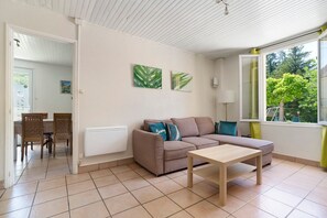A living room with a sofa, a table, and a window with a view of greenery. - Holiday Home La Mailleraie - L'étang with Shared Pool, Private Terrace & Wi-Fi (Valojoulx)