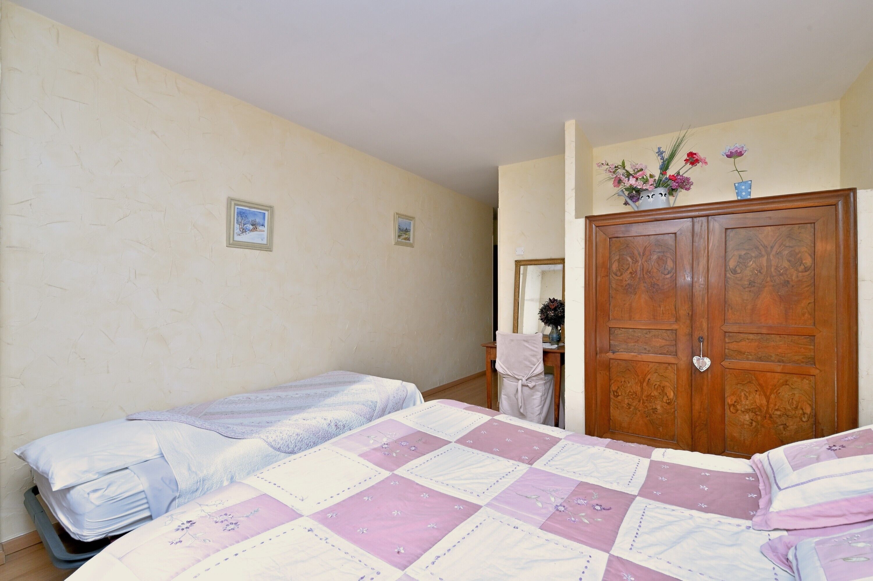 A bedroom with two beds, a wooden wardrobe, and a small table with a vase of flowers.