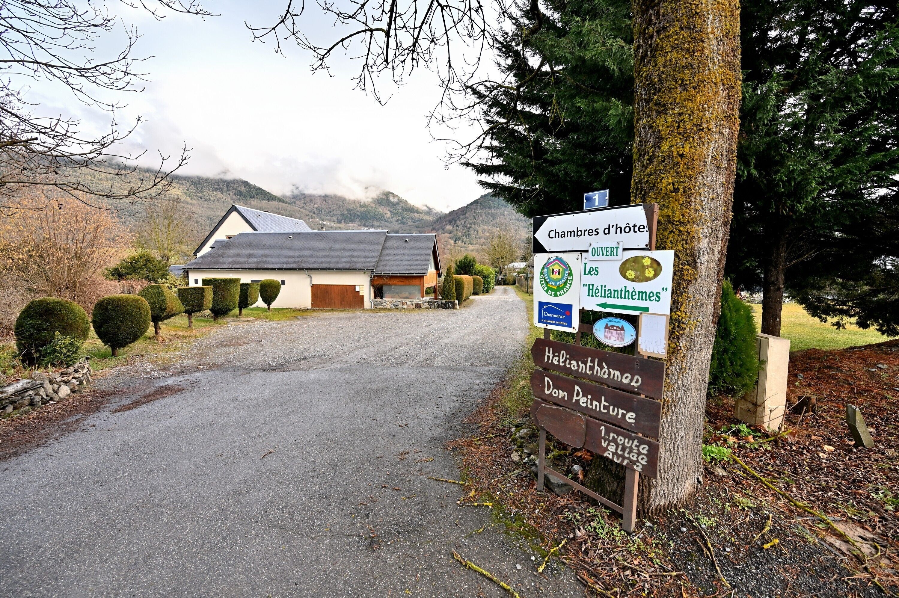 A rural road leading to a house with a sign indicating 'Chambres d'hôtes' and 'Hélianthemes' nearby.